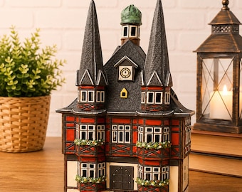 May include: A ceramic house figurine featuring a half-timbered design with red and cream walls, two tall dark gray spires, a central clock tower, and a green dome. The structure includes multiple windows with flower boxes and a front arched door.