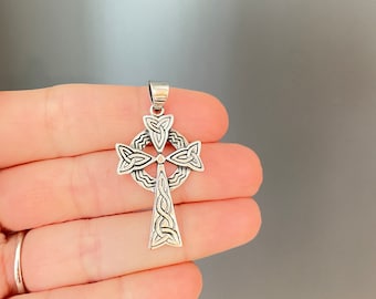 May include: A silver Celtic cross pendant with detailed knotwork. The cross has a circular feature where the arms meet and a loop for a chain. The pendant is held in a hand against a grey background.