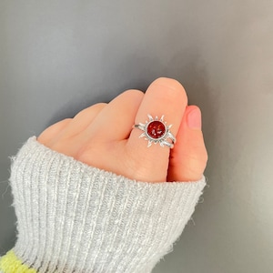 K&ouml;nnte beinhalten: Ein silberner Ring mit einem Sonnenmuster und einem zentralen, runden, rot-orangen Stein. Der Ring hat ein Doppelband und wird am Finger getragen. Das Design des Rings erinnert an ein himmlisches Thema, passend f&uuml;r Alltag oder besondere Anl&auml;sse.
