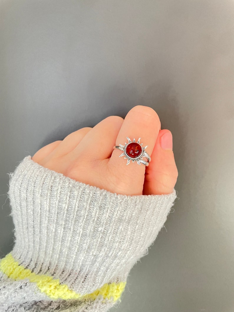 K&ouml;nnte beinhalten: Ein silberner Ring mit einem Sonnenmuster und einem zentralen, runden, rot-orangen Stein. Der Ring hat ein Doppelband und wird am Finger getragen. Das Design des Rings erinnert an ein himmlisches Thema, passend f&uuml;r Alltag oder besondere Anl&auml;sse.