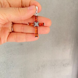 Puede incluir: Un colgante de cruz de plata con un centro de &aacute;mbar marr&oacute;n. La cruz est&aacute; decorada con peque&ntilde;os detalles plateados.