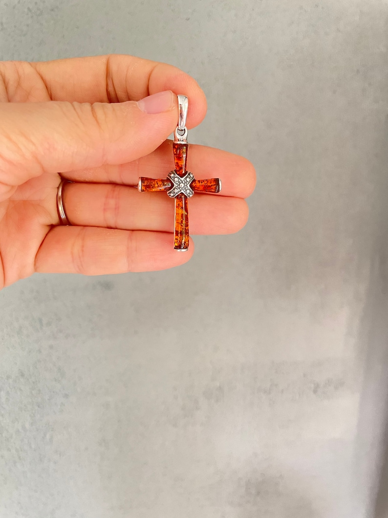 Puede incluir: Un colgante de cruz de plata con un centro de &aacute;mbar marr&oacute;n. La cruz est&aacute; decorada con peque&ntilde;os detalles plateados.