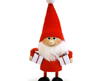 Puede incluir: Un gnomo navideño rojo y blanco sosteniendo dos cajas de regalo blancas. El gnomo tiene una barba blanca y una cara sonriente.