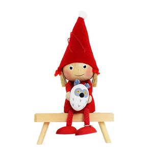 May include: A red felt gnome doll with a white hat sits on a wooden bench. The gnome is holding a white felt owl with gray eyes.