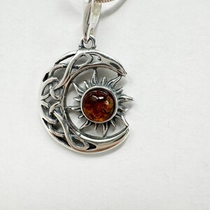 May include: A silver pendant necklace featuring a crescent moon design with Celtic knotwork and a sunburst. The sun has a round, amber-colored center. The pendant hangs from a delicate silver chain.