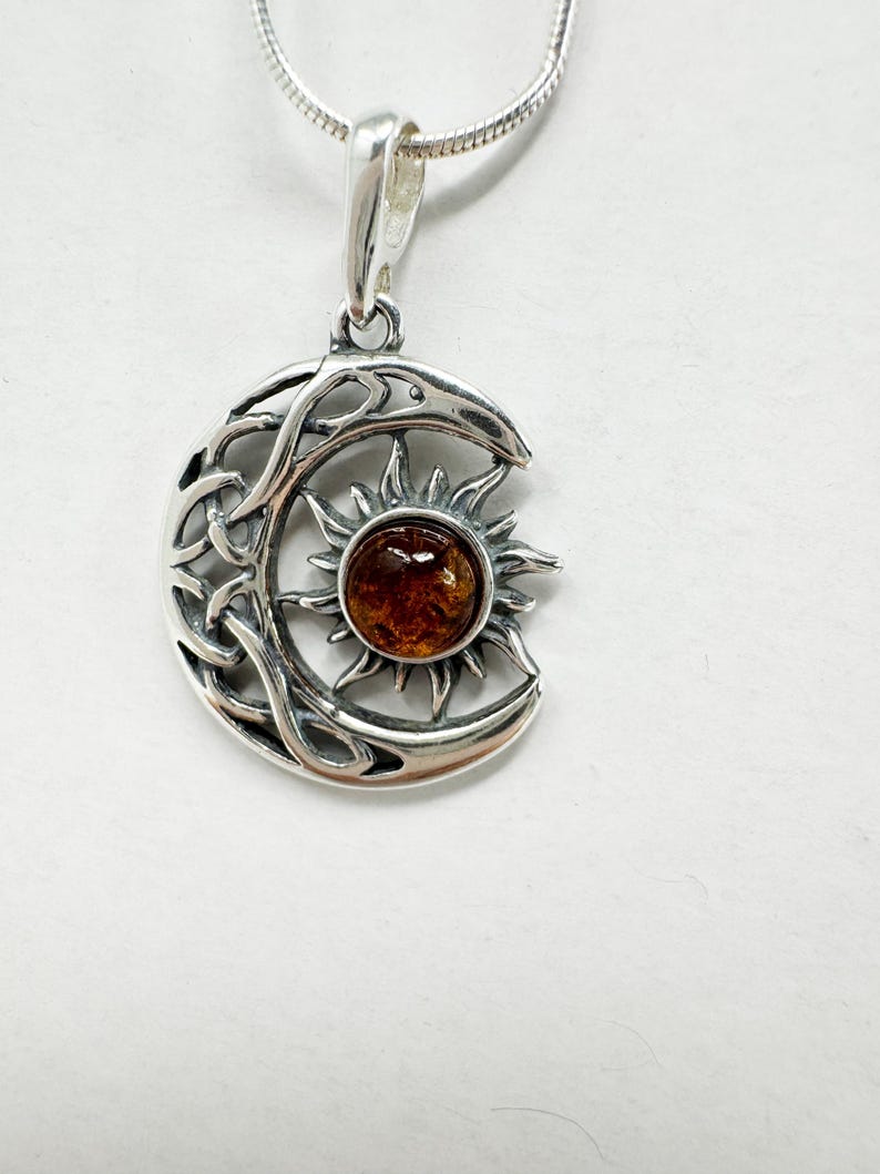May include: A silver pendant necklace featuring a crescent moon design with Celtic knotwork and a sunburst. The sun has a round, amber-colored center. The pendant hangs from a delicate silver chain.