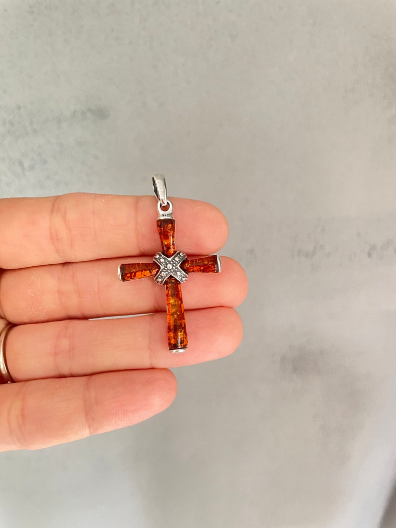 Puede incluir: Un colgante de cruz de plata con una incrustaci&oacute;n de piedra &aacute;mbar marr&oacute;n y un peque&ntilde;o acento de diamante.