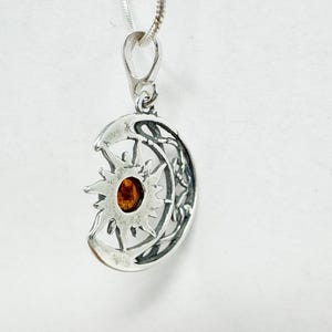 May include: A silver pendant necklace featuring a sun and moon design. The pendant has a detailed, openwork design with a central amber-colored stone. The necklace chain is a thin, silver cable.