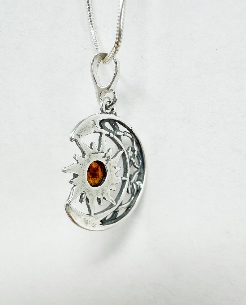 May include: A silver pendant necklace featuring a sun and moon design. The pendant has a detailed, openwork design with a central amber-colored stone. The necklace chain is a thin, silver cable.