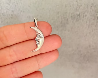 May include: A silver crescent moon pendant with a face on it. The pendant is hanging from a silver chain.