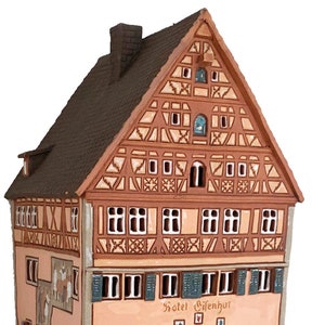 May include: A ceramic model of a traditional German building with a brown roof, white trim, and a sign that reads "Hotel Gifenhur". The building has a half-timbered facade and is decorated with intricate details.