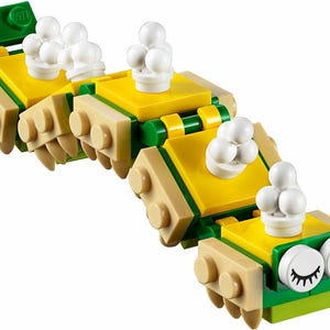 Könnte beinhalten: Ein Lego-Raupenspielzeug aus gelben, grünen und beigen Blöcken. Das Spielzeug hat weiße, runde Teile oben und weiße Augen mit schwarzen Wimpern vorne.