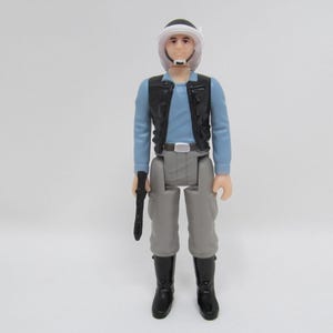 May include: A black and gray action figure wearing a white helmet, a blue shirt, a black vest, and gray pants. The figure is holding a black gun.