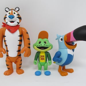 May include: Three colorful cartoon character figurines: a tiger, a frog, and a toucan. The tiger is orange with black stripes and a red bandana. The frog is green with a yellow shirt. The toucan is blue with a large, multi-colored beak.