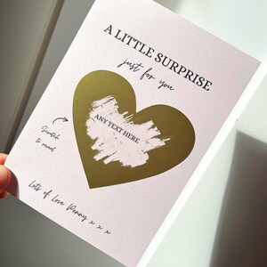 Puede incluir: Una tarjeta rosa con un coraz&oacute;n de l&aacute;mina dorada y el texto "A LITTLE SURPRISE just for you". La tarjeta tiene un &aacute;rea para rascar con las palabras "ANY TEXT HERE" y "Scratch to reveal".