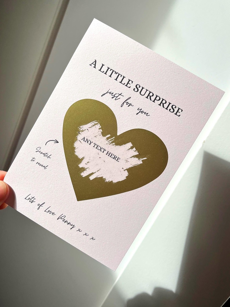 Puede incluir: Una tarjeta rosa con un coraz&oacute;n de l&aacute;mina dorada y el texto "A LITTLE SURPRISE just for you". La tarjeta tiene un &aacute;rea para rascar con las palabras "ANY TEXT HERE" y "Scratch to reveal".