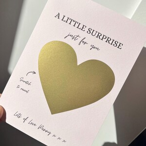 Puede incluir: Tarjeta rosa claro para rascar con un coraz&oacute;n dorado en el centro. El texto negro dice "A LITTLE SURPRISE just for you". Debajo, se lee "Scratch to reveal" y "Lots of Love Penny x x x."