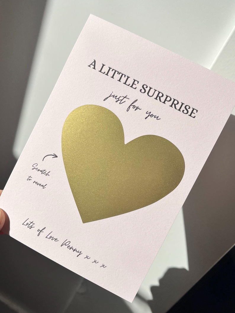 Puede incluir: Tarjeta rosa claro para rascar con un coraz&oacute;n dorado en el centro. El texto negro dice "A LITTLE SURPRISE just for you". Debajo, se lee "Scratch to reveal" y "Lots of Love Penny x x x."