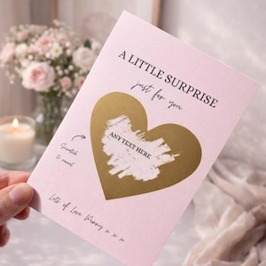 Puede incluir: Una tarjeta rosa para rascar con un coraz&oacute;n dorado y el texto "A LITTLE SURPRISE just for you". La tarjeta tiene el texto "Scratch to reveal" y "ANY TEXT HERE". La tarjeta est&aacute; sujeta en una mano.
