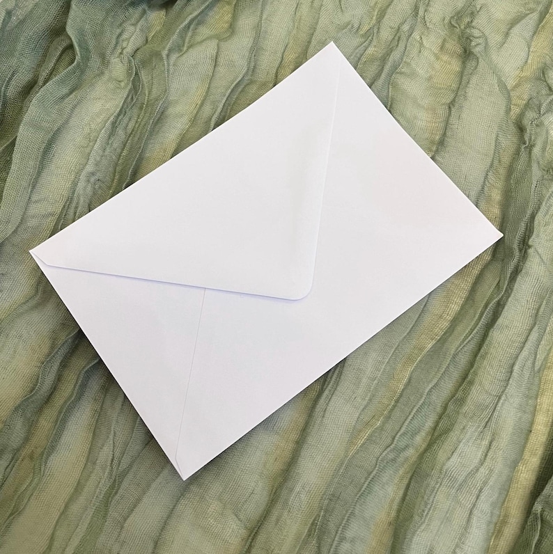 Puede incluir: Un sobre blanco con solapa triangular, sobre una tela verde oliva texturizada. El sobre es limpio y sencillo, con un dise&ntilde;o cl&aacute;sico. La tela tiene un aspecto arrugado.