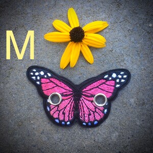 Peut inclure: Un patch en forme de papillon rose avec des contours noirs et des accents blancs. Le patch a deux &oelig;illets m&eacute;talliques et la lettre "M" est visible en arri&egrave;re-plan.