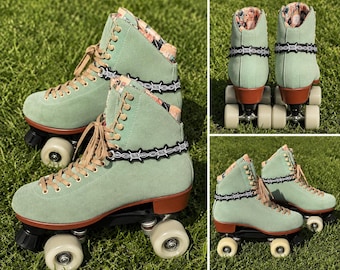 May include: A pair of mint green roller skates with brown laces and wheels. The skates have a floral pattern on the collar and a black barbed wire design around the ankle. The wheels are off-white and the base is black.