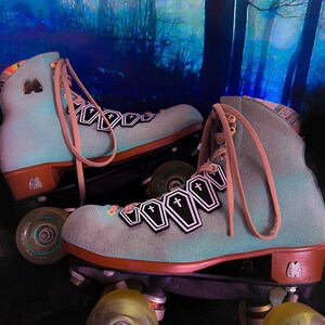 May include: A pair of teal blue roller skates with black coffin-shaped patches and a white cross on each patch. The skates have brown leather straps and wheels with a teal blue rim.