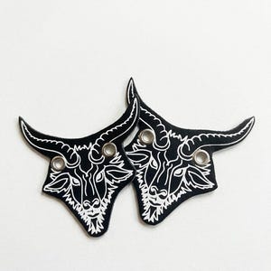 Könnte beinhalten: Zwei schwarze Lederaufnäher, jeweils mit einer weißen Illustration eines Stierkopfes mit Hörnern. Das Stierkopfdesign ist mit weißen Linien detailliert. Jeder Aufnäher hat eine silberne Öse zur Befestigung. Auf weißem Hintergrund.