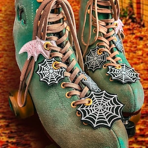 May include: A pair of green roller skates with black and white spiderweb charms and a pink bat charm. The skates have brown laces and gold accents.