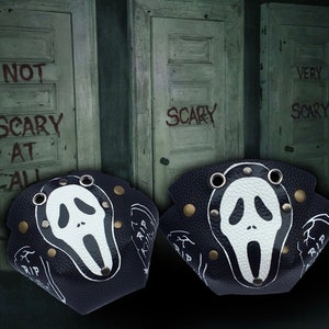 May include: Two black wrist cuffs with a white Ghostface mask design, brass studs, and silver eyelets. The cuffs have a textured appearance and a RIP tombstone graphic. The background features three weathered doors with the words "NOT SCARY AT ALL" and "SCARY" written on them.