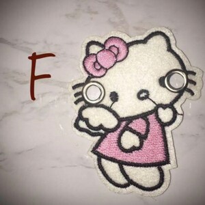 May include: White embroidered Hello Kitty patch with pink bow, pink dress, and black outlines. The patch has two silver eyelets.