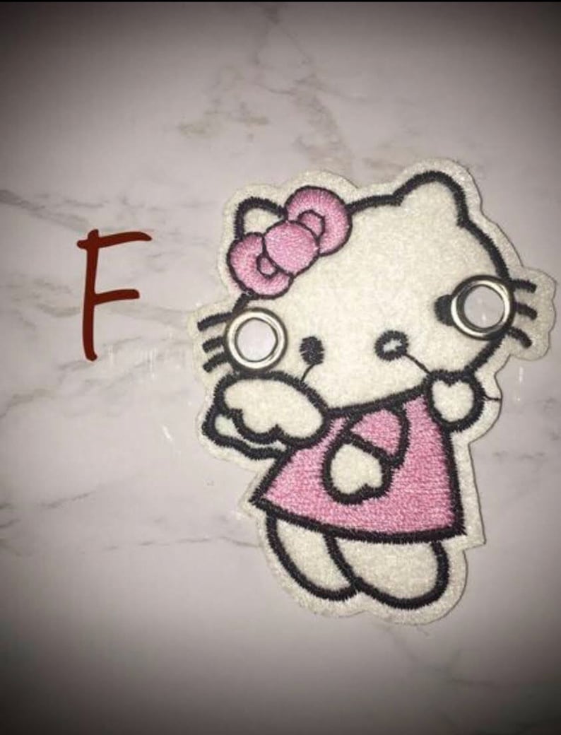 May include: White embroidered Hello Kitty patch with pink bow, pink dress, and black outlines. The patch has two silver eyelets.