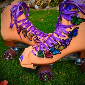 Puede incluir: Un par de patines de ruedas de color canela con cordones morados y parches de mariposas de colores. Los patines tienen ruedas negras y un marco de metal negro.