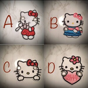 May include: Four embroidered iron-on patches featuring Hello Kitty in different poses. The patches are white with black outlines and feature different colors and details. The first patch shows Hello Kitty holding a camera, the second shows Hello Kitty wearing a blue dress, the third shows Hello Kitty's face, and the fourth shows Hello Kitty holding a pink heart with the text "Hello Kitty".