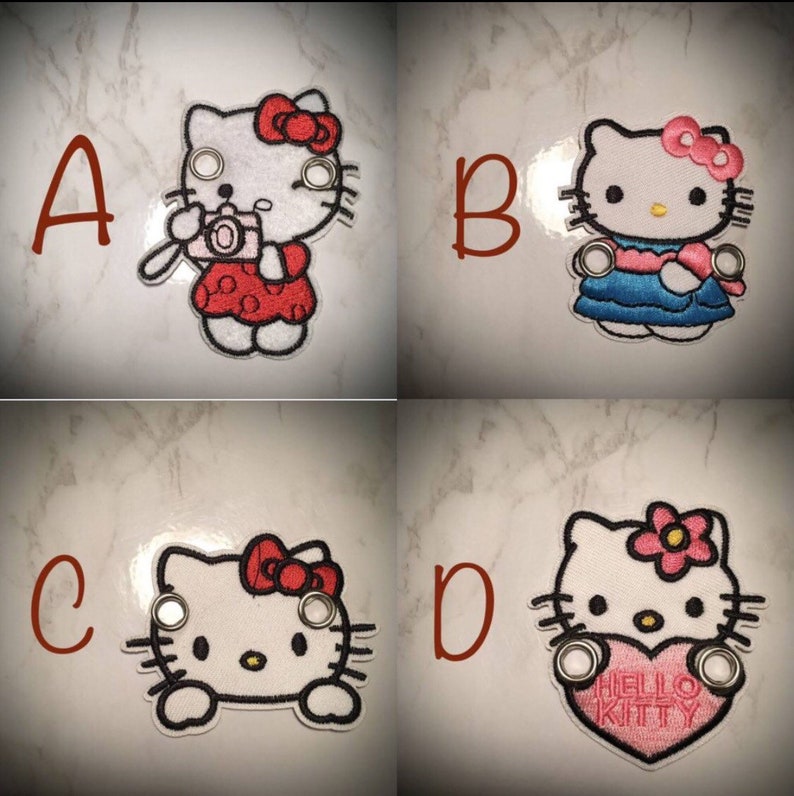 May include: Four embroidered iron-on patches featuring Hello Kitty in different poses. The patches are white with black outlines and feature different colors and details. The first patch shows Hello Kitty holding a camera, the second shows Hello Kitty wearing a blue dress, the third shows Hello Kitty's face, and the fourth shows Hello Kitty holding a pink heart with the text "Hello Kitty".