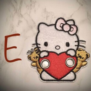 May include: White embroidered patch with a cartoon Hello Kitty holding a red heart and two gold teddy bears. The patch has two silver eyelets.