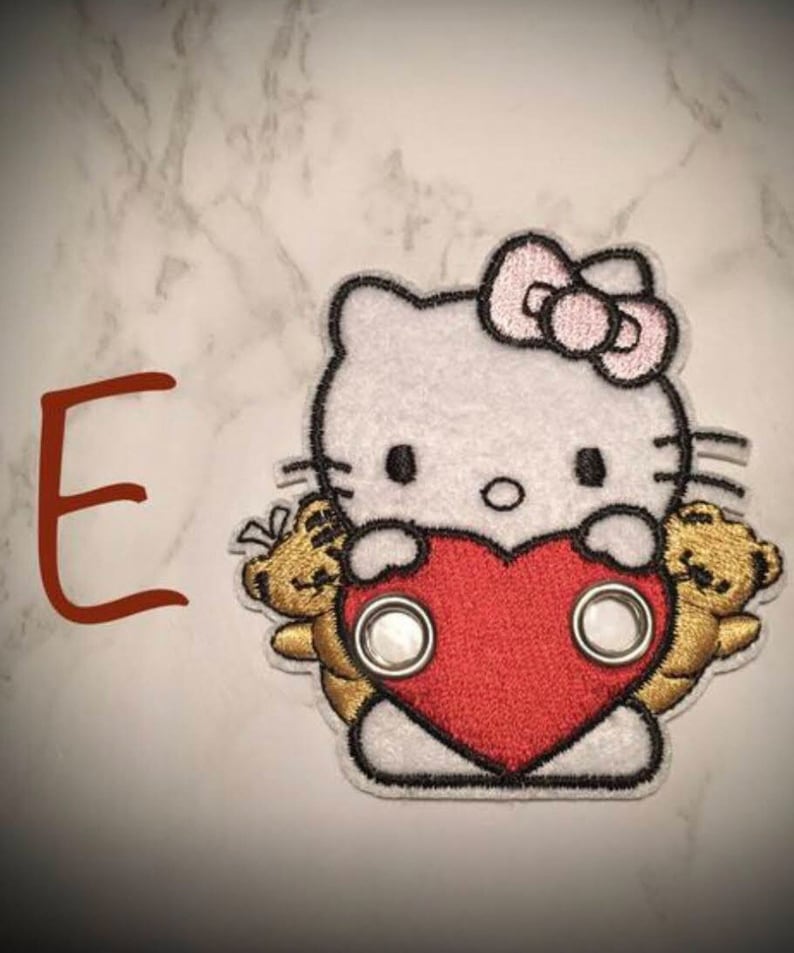 May include: White embroidered patch with a cartoon Hello Kitty holding a red heart and two gold teddy bears. The patch has two silver eyelets.