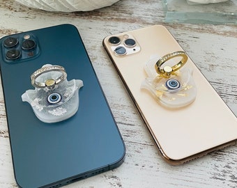 May include: Two iPhone cases with a clear, hamsa hand design with a blue evil eye. One case is teal and the other is gold.