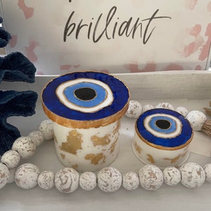 May include: Two white ceramic containers with gold accents and blue evil eye designs. The larger container is taller and has a more detailed design. The smaller container is shorter and has a simpler design. Both containers are sitting on a white surface with white beads.