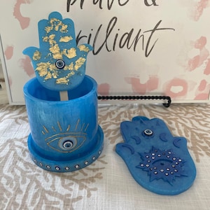 May include: Blue ceramic Hamsa hand incense holder with a gold evil eye and gold leaf accents. The holder is open and the lid is a separate Hamsa hand with a blue evil eye and a starburst design.