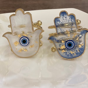 May include: Two gold hamsa hand napkin rings with blue evil eye details. One ring is white with gold flecks and the other is blue with gold flecks.