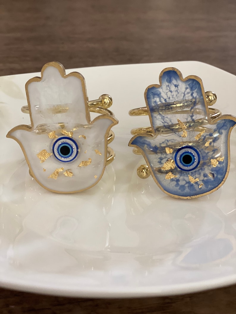 May include: Two gold hamsa hand napkin rings with blue evil eye details. One ring is white with gold flecks and the other is blue with gold flecks.