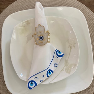 May include: White napkin ring featuring a gold hamsa hand design and a blue evil eye. The ring is placed on a white napkin with a blue evil eye border. The napkin is on a white plate with a floral design.