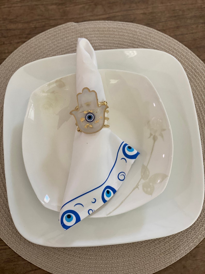 May include: White napkin ring featuring a gold hamsa hand design and a blue evil eye. The ring is placed on a white napkin with a blue evil eye border. The napkin is on a white plate with a floral design.