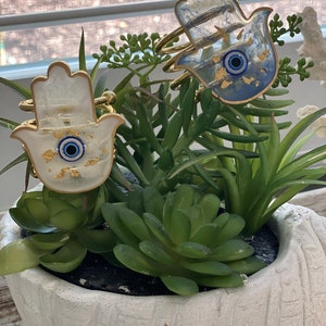 May include: Two hamsa hand charms with gold accents and blue evil eye details. The charms are made of white and blue resin and are sitting in a white planter with green succulents.