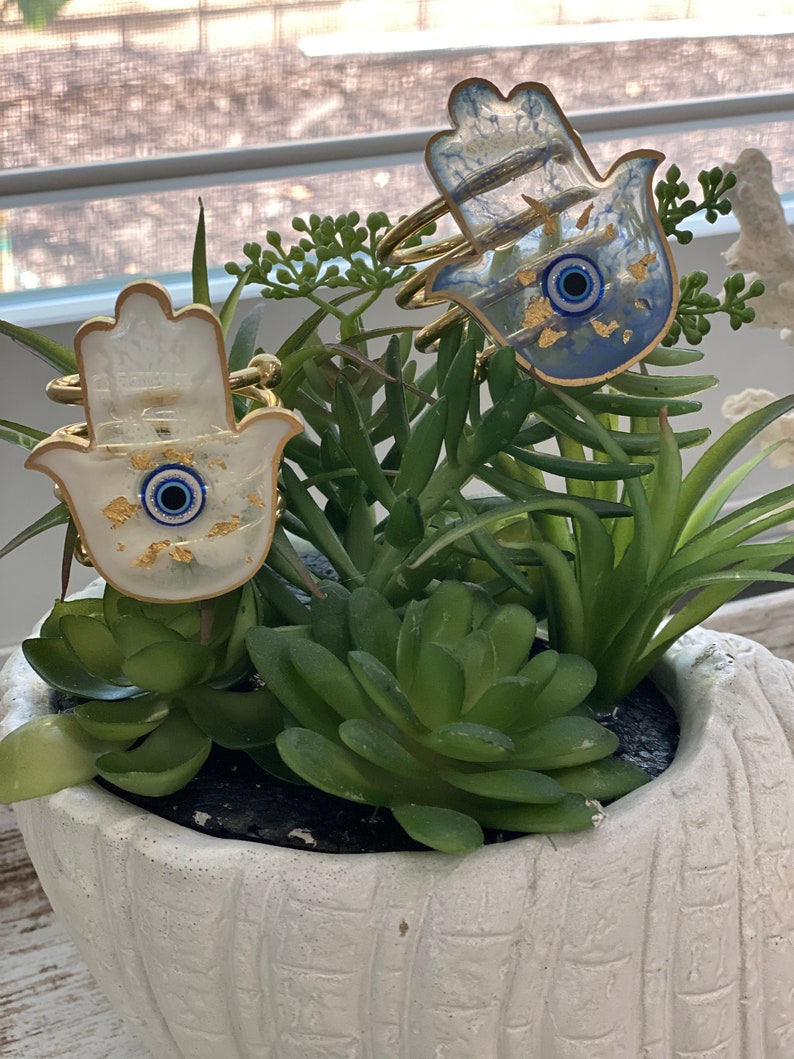May include: Two hamsa hand charms with gold accents and blue evil eye details. The charms are made of white and blue resin and are sitting in a white planter with green succulents.