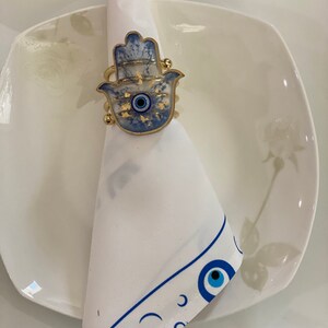 May include: A white napkin with a blue and white eye design border. A gold hamsa hand with a blue eye charm is used as a napkin ring.