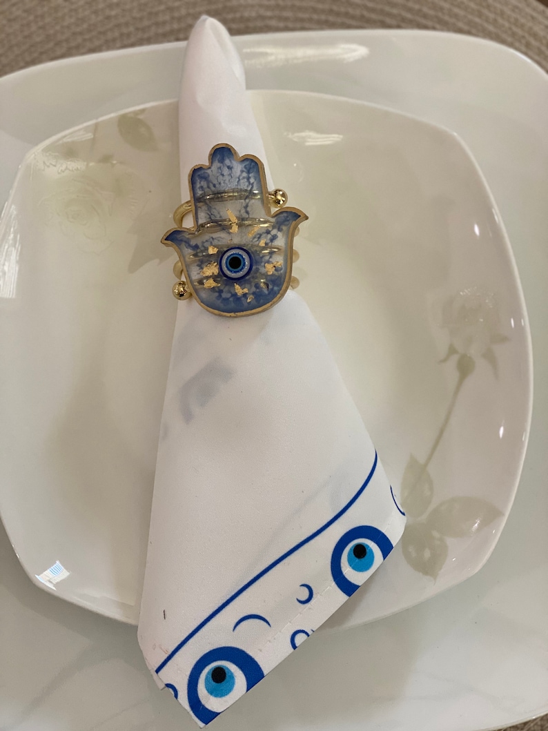 May include: A white napkin with a blue and white eye design border. A gold hamsa hand with a blue eye charm is used as a napkin ring.