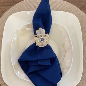 May include: A blue napkin ring with a white hamsa hand design and a blue evil eye charm. The ring is on a white plate with a beige placemat.