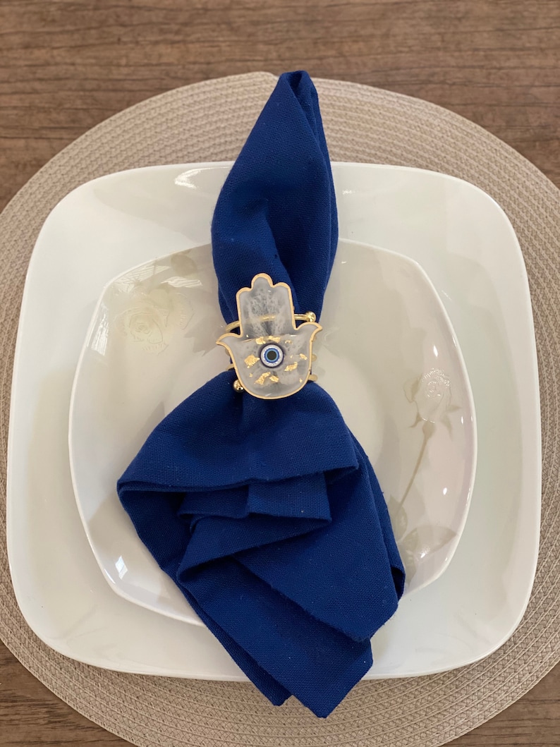 May include: A blue napkin ring with a white hamsa hand design and a blue evil eye charm. The ring is on a white plate with a beige placemat.